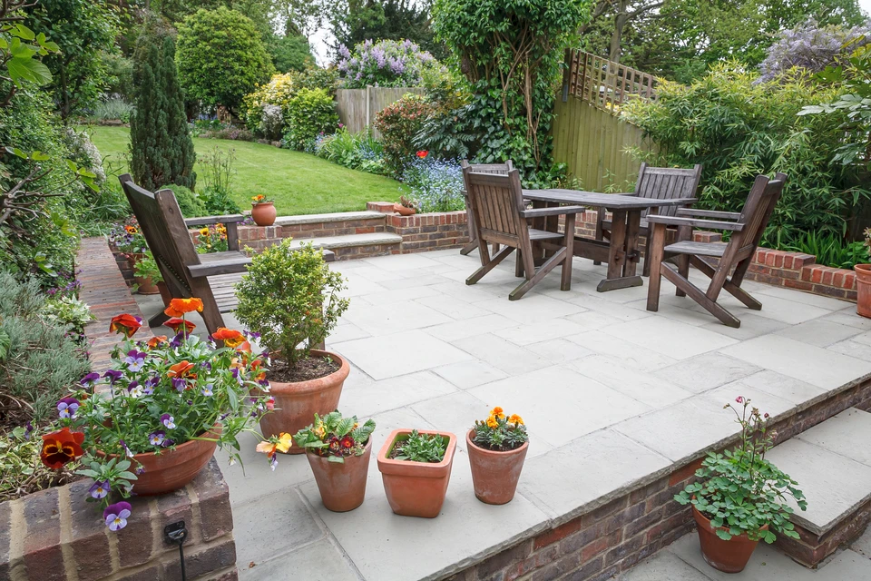 Terrasse extérieure aménagée avec salon de jardin en bois, jardin paysager en arrière-plan, bordures végétalisées et pots de fleurs colorées