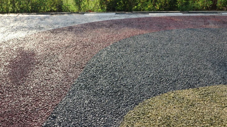 Photographie professionnelle en couleur montrant une allée extérieure récemment aménagée en stabilisé calcaire ou grave compactée, bordée de caniveaux de drainage en béton. Au premier plan, une pelleteuse compacte sur pneus jaune et noire effectue les finitions de nivellement. En arrière-plan, un terrain avec travaux de terrassement en cours, muret de soutènement en pierre, et prairies verdoyantes. L'image doit capturer l'expertise en aménagement extérieur, préparation du sol et drainage pour cours et terrains, représentative d'un chantier VRD professionnel en milieu rural.