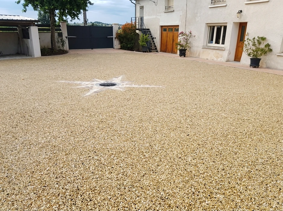 Cour extérieure pavée en résine drainante ton beige/ocre, avec motif décoratif étoile à 8 branches en granulats noirs et blancs centré sur un regard de sol