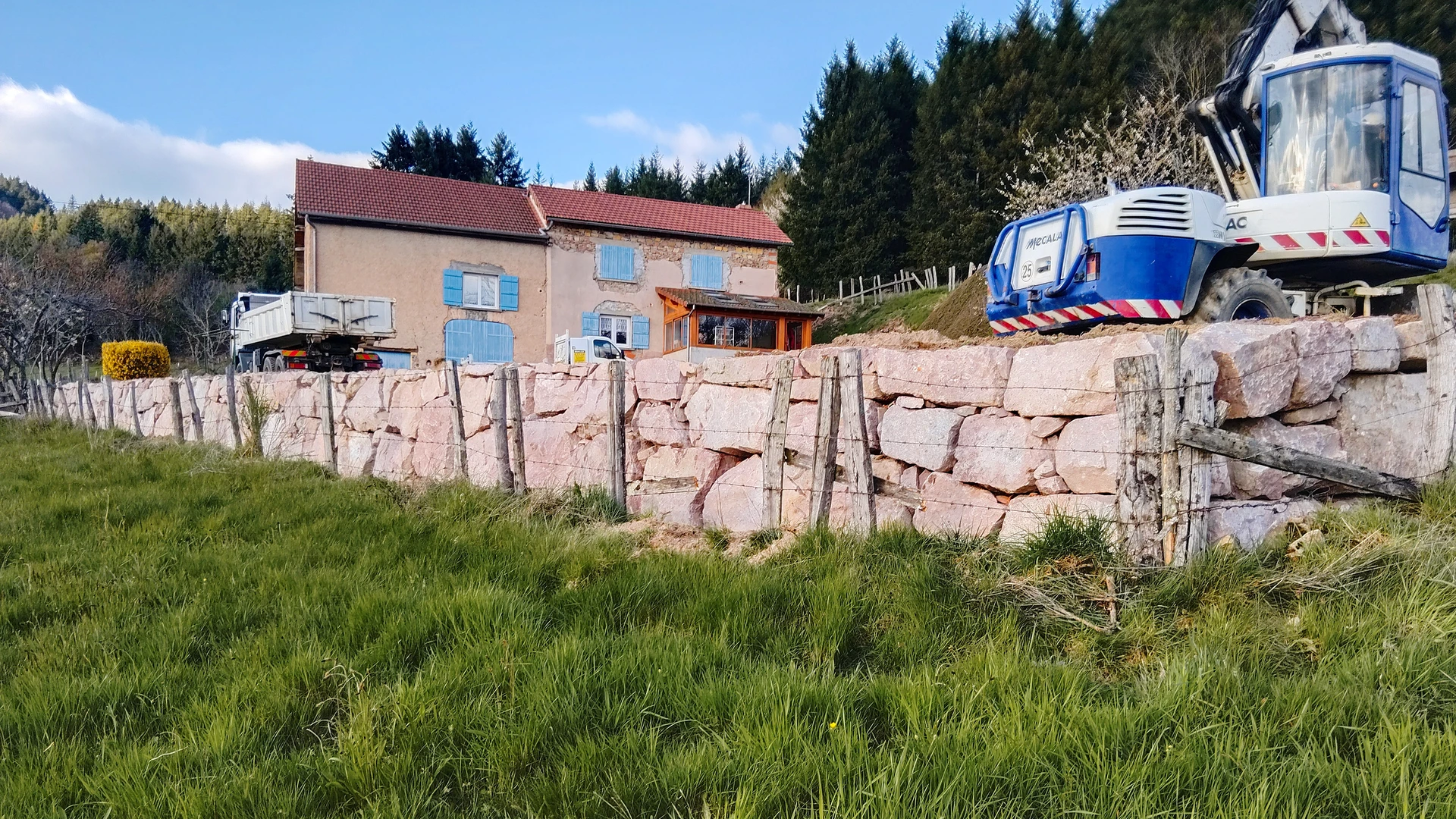 Chantier d'enrochement professionnel : une pelle hydraulique sur pneus équipée d'un godet de fouille positionne de gros blocs de pierre calcaire ou granite sur une berge ou un terrain en pente. Les roches sont empilées de manière structurée pour créer une stabilisation de talus. On aperçoit une excavatrice en action, des remblais de terre en arrière-plan, et une zone de travail nettement délimitée. Conditions de jour clairs, ciel bleu ou dégagé, image de qualité professionnelle montrant l'expertise technique de l'entreprise.